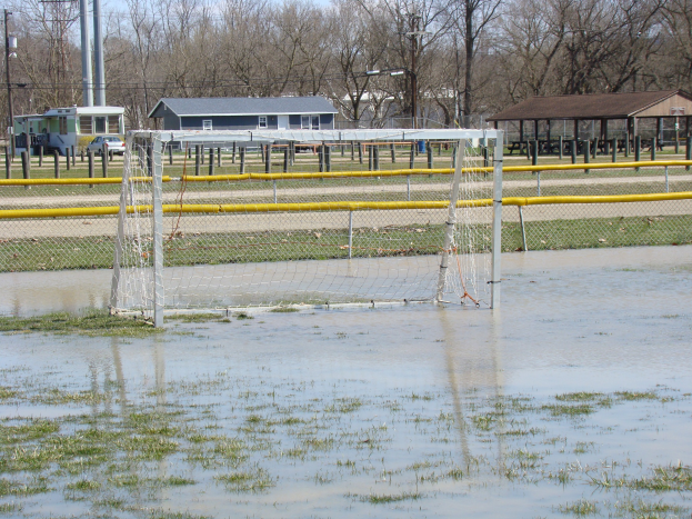 Ein Fußballtor steht in der Mitte eines überfluteten Feldes, umgeben von Gras und Wasser, vor einer Kulisse aus Schuppen, Pfählen, Bäumen, Fahrzeugen und einem klaren blauen Himmel.
