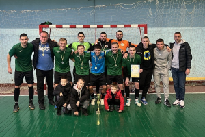Eine Gruppe von Männern in Medaillen, einer hält einen Pokal, steht auf einem Futsal-Platz mit einem Torpfosten und einer Wand im Hintergrund.