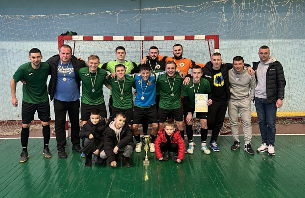 Eine Gruppe von Männern in Medaillen, einer hält einen Pokal, steht auf einem Futsal-Platz mit einem Torpfosten und einer Wand im Hintergrund.