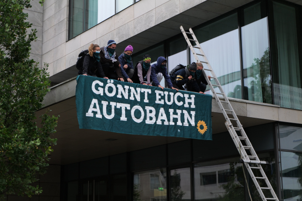 Gruppe von Menschen auf einem Gebäudedach mit einer Fahne, mit einem Baum links und Glasfenstern im Hintergrund, die scheinbar protestieren und die deutsche Regierung unterstützen.