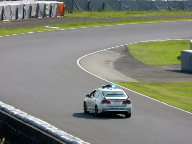 Polizeifahrzeug auf einer Rennstrecke mit Gras auf beiden Seiten und Zuschauern und Zelten im Hintergrund.