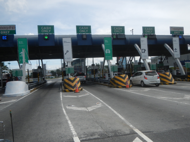 Eine Mautstation an der Seite einer Straße mit darunter fahrenden Autos, umgeben von Verkehrskegeln, Absperrpfosten, Schildern, einem Tor, Laternenmasten, Bäumen, Gebäuden und einem bewölkten Himmel.