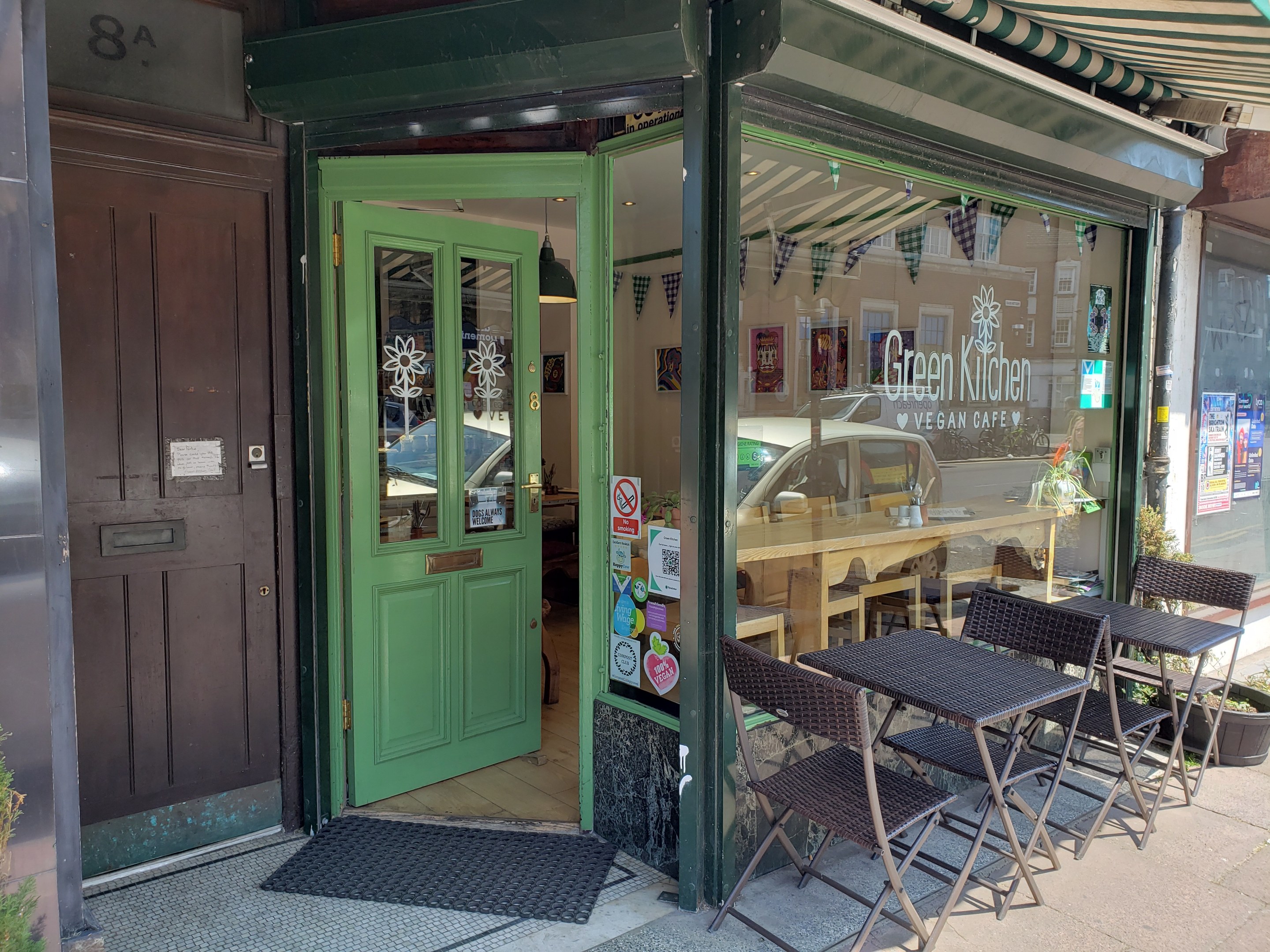 Außensitzbereich des Green Kitchen Vegan Cafe mit Tischen, Stühlen, Pflanzen, einer Fußmatte und einer Glastür mit Postern, die in einen Innenraum mit Tischen, Stühlen und Deckenleuchten führen.