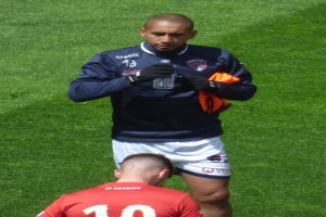 Ein Mann in einem Sportoutfit steht auf einem grünen Feld und hält in einer Hand eine Flasche und in der anderen ein oranges Tuch.