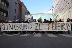 Demonstranten auf einer Straße halten ein Transparent mit der Aufschrift "Kein Grund zu Feiern" gegen die deutschen Sparmaßnahmen, mit Gebäuden, einem Schild, einem weiteren Transparent, einem Kran und einem bewölkten Himmel im Hintergrund.