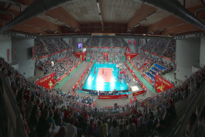 Eine große Halle voller Zuschauer bei einem Volleyballspiel mit Bannern, Bildschirmen und Deckenbeleuchtung sowie Geländern unten.