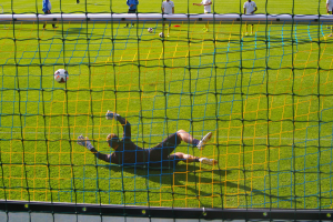 Ein Fussballer hält den Ball vor dem Tor, mit einem Netz im Vordergrund und einigen Personen im Hintergrund, während mehrere Bälle auf dem Boden verstreut sind.