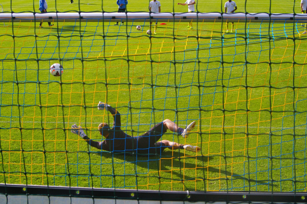 Ein Fussballer hält den Ball vor dem Tor, mit einem Netz im Vordergrund und einigen Personen im Hintergrund, während mehrere Bälle auf dem Boden verstreut sind.