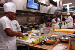 Eine Gruppe von Köchen in einer Küche beim Zubereiten von Speisen auf einem Schneidbrett, wobei ein Koch im Vordergrund einen weißen Kittel und eine Haube trägt und ein Messer hält und von verschiedenen Utensilien, Schüsseln, Gemüse, einem Mobiltelefon, Flaschen und anderen Gegenständen umgeben ist, während im Hintergrund ein Fernseher zu sehen ist.