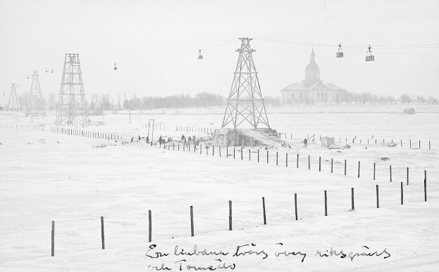 Ein Schwarz-Weiß-Foto einer Sesselbahn in einer verschneiten Wiese, umgeben von Pfählen, Drähten, Bäumen und einem Gebäude im Hintergrund, mit Text unten.