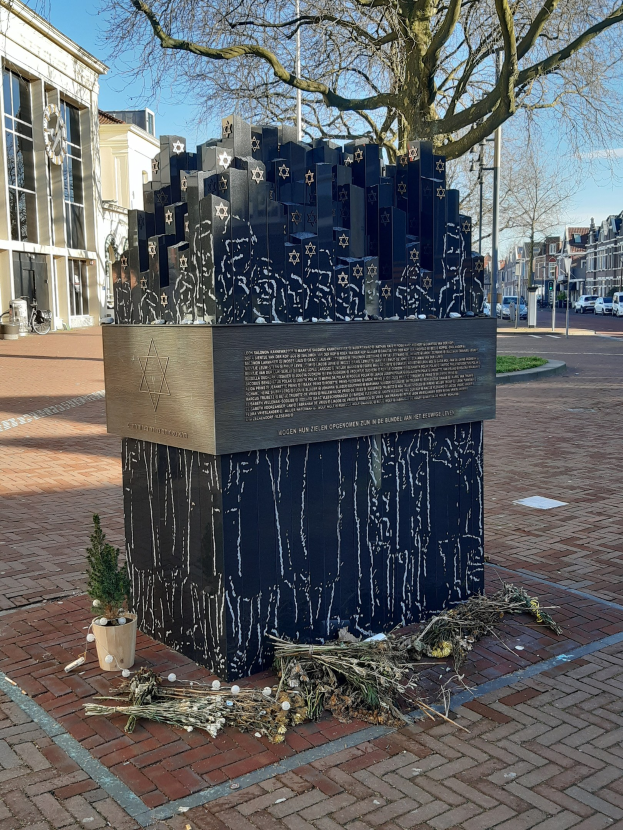 Denkmal in einem Stadtplatz für Holocaust-Opfer, mit einem Baum davor, einem Topfgewächs, verstreuten trockenen Blättern und Gebäuden im Hintergrund.