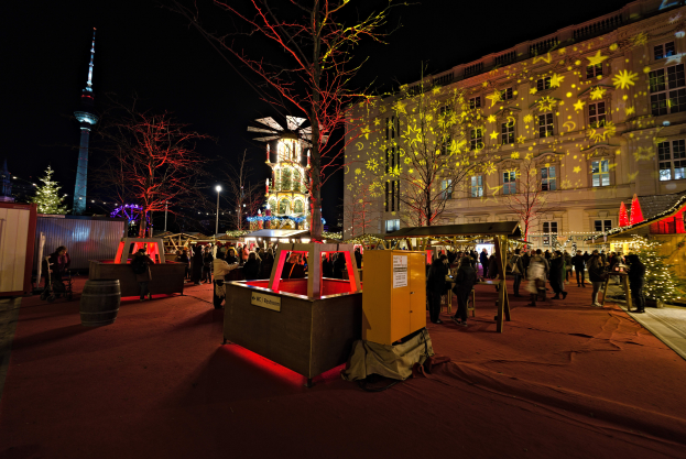 Ein lebendiger Weihnachtsmarkt in Berlin, Deutschland, mit Menschen um beleuchtete Stände, Bäume, Gebäude, Laternenpfähle und einen Turm unter einem dunklen Himmel.
