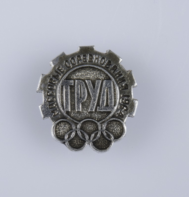 Ein silbernes Abzeichen mit der Aufschrift "Olympische Spiele", vor einem weißen Hintergrund.