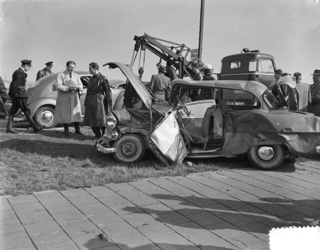Schwarz-weißes Bild eines beschädigten Autos am Straßenrand mit mehreren Menschen drumherum und einem Pfahl im Hintergrund.