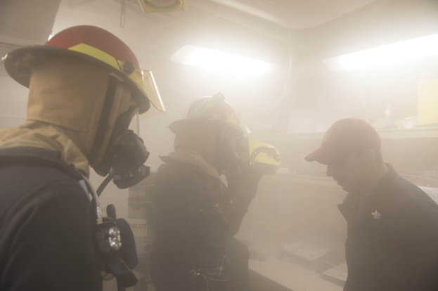 Feuerwehrleute in einem raucherfüllten Raum während einer Übung, einer hält eine Kamera, mit Hintergrundbeleuchtung und -ausrüstung.