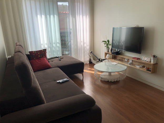 Ein Wohnzimmer mit einer Couch, einem Flachbildfernseher an der Wand, einem Tisch mit verschiedenen Gegenständen vor der Couch und einem Fenster mit Vorhängen, das Gebäude und Himmel draußen zeigt.