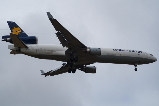 Lufthansa Cargo Boeing 777-300ER im Flug mit sichtbarem Text auf dem Rumpf, ausgefahrenen Tragflächen, aktive Triebwerke, vor einem Himmel mit weißen Wolken.