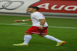 Ein Mann in weißem Hemd und roter Hose läuft auf einem Fußballfeld, trägt Fußballschuhe und hat ein Schild im Hintergrund.