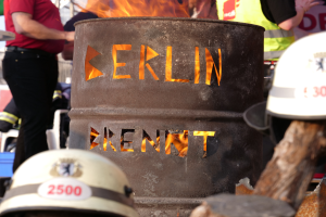 Eine Gruppe von Menschen um ein Feuerhydrant mit der Aufschrift "Berlin" herumstehend mit einem brennenden Fass im Vordergrund, Helme unten und ein hölzernes Objekt auf der rechten Seite.