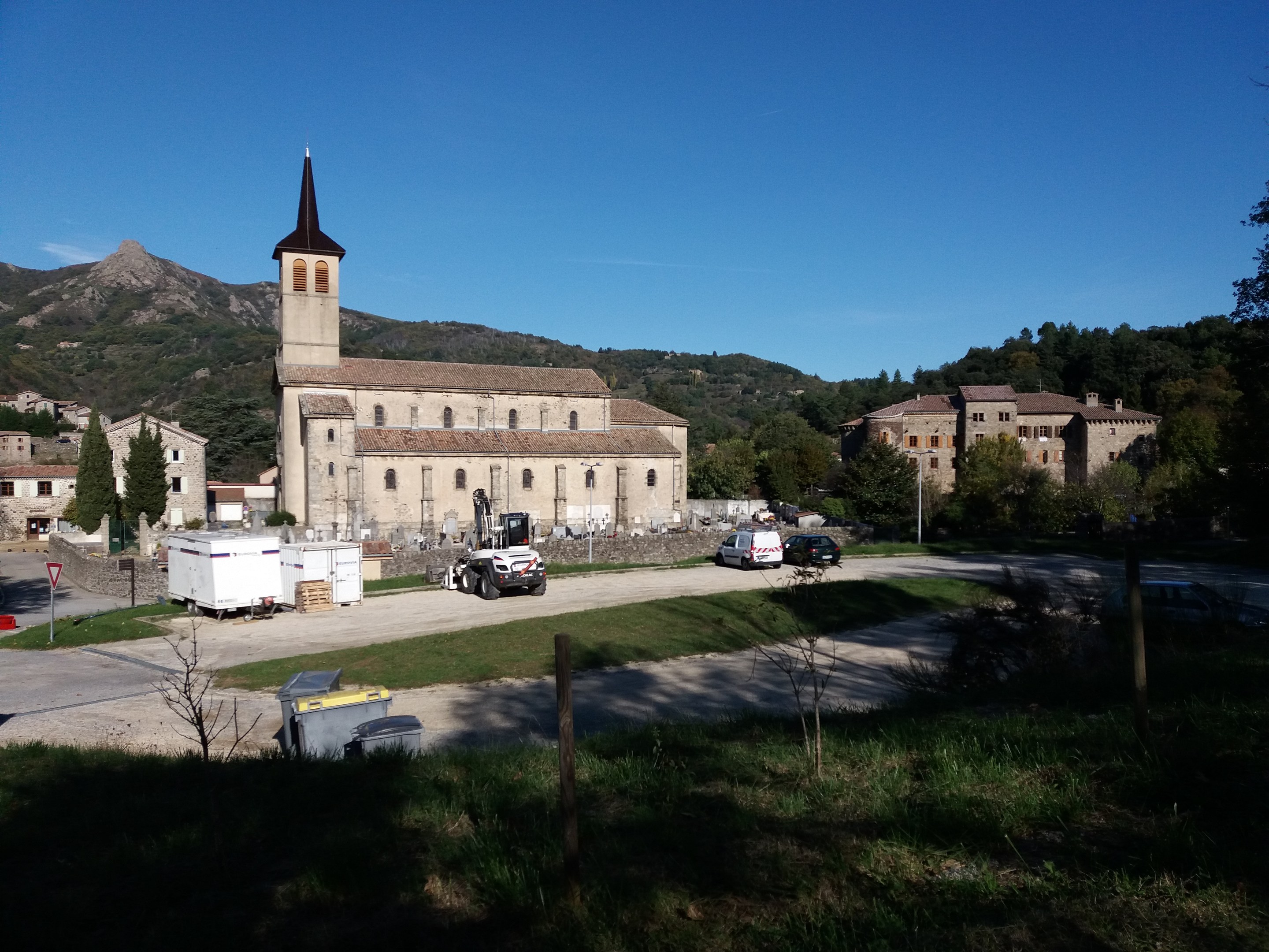 Luftaufnahme einer Kleinstadt mit einer zentralen Kirche, umgeben von Gebäuden, Bäumen, Straßen mit Fahrzeugen, Gras, Strommasten und Schildern, vor einem Hügel- und einem klaren blauen Himmel.