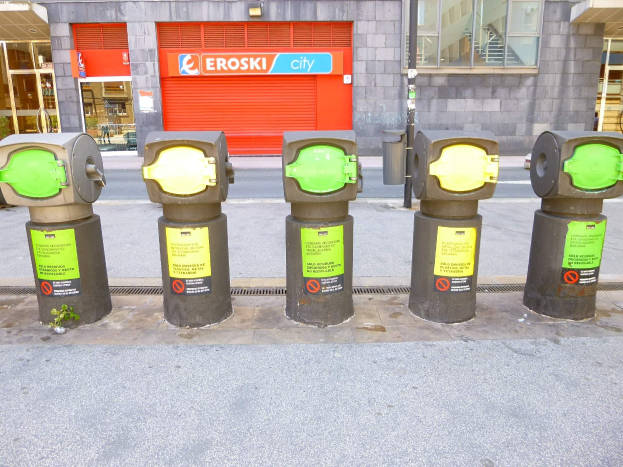 Reihe von Parkautomaten entlang einer Straße mit einem Gebäude, das Glasfenster und eine Rolllade mit Text sowie einen nahen Müllcontainer zeigt.