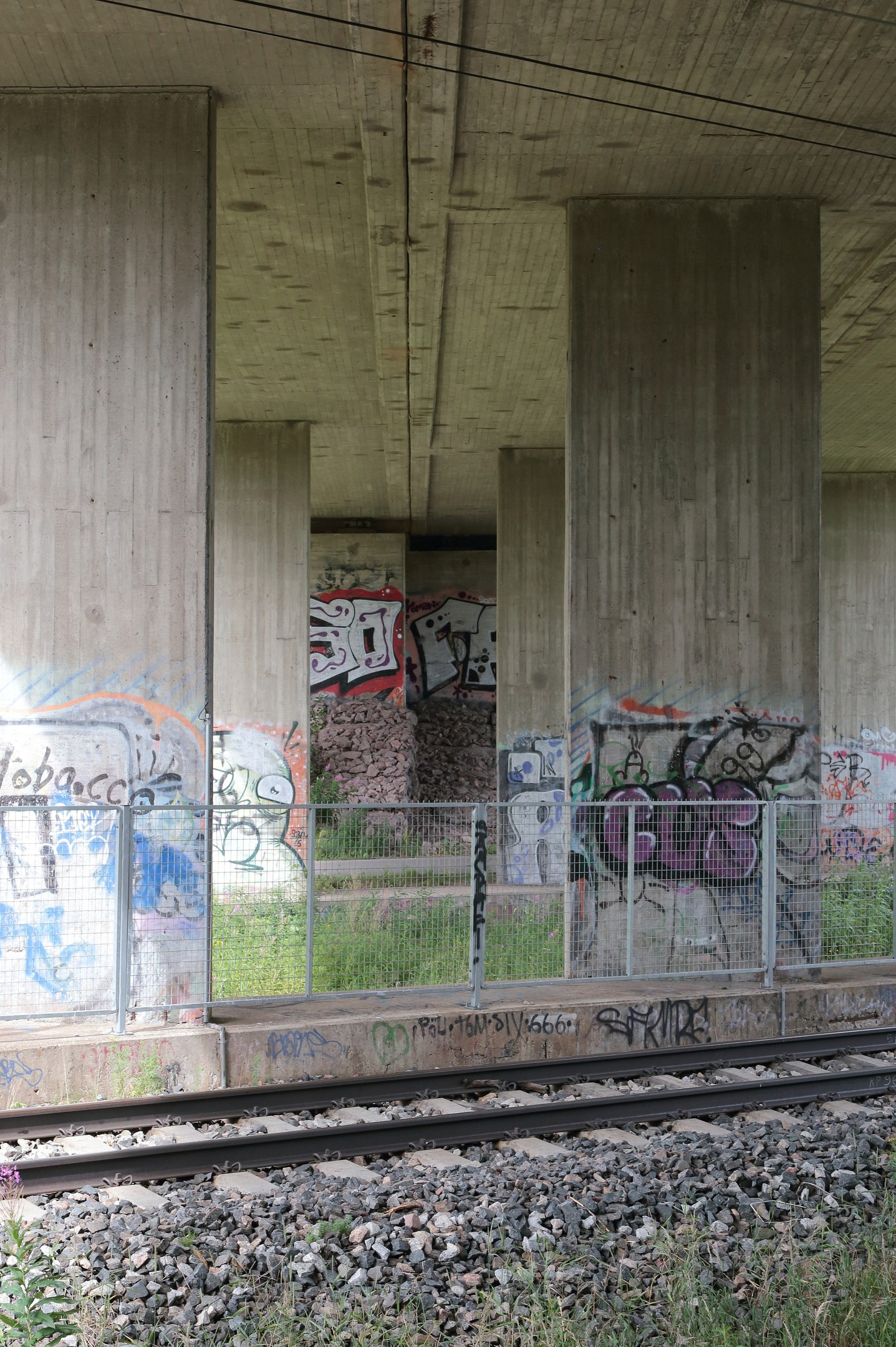 Eisenbahnschiene mit Graffiti an einer Betonwand, umgeben von Steinen, Gras, einem Zaun, Säulen, Oberleitungen und einem Dach.