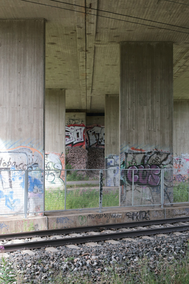 Eisenbahnschiene mit Graffiti an einer Betonwand, umgeben von Steinen, Gras, einem Zaun, Säulen, Oberleitungen und einem Dach.