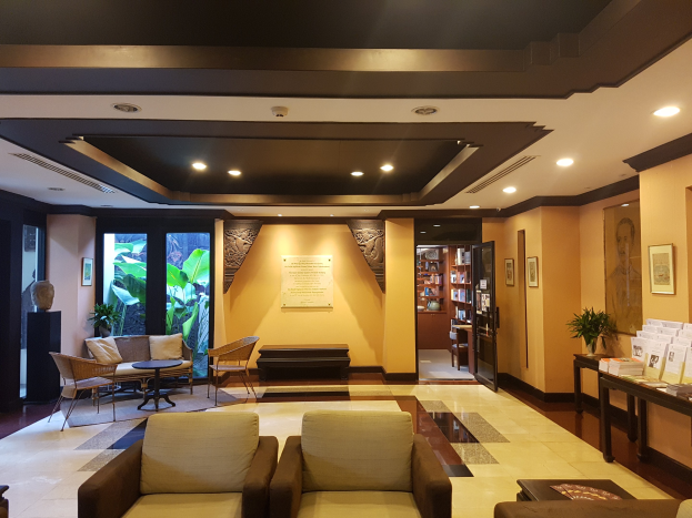 Ein geräumiger Hotellobby mit Sitzbereichen, Dekorationspflanzen, Bücherregalen, Leuchten, Wandkunst und Seitentisch-Dekoration, beleuchtet von natürlichem Licht durch Fenster und Türen.