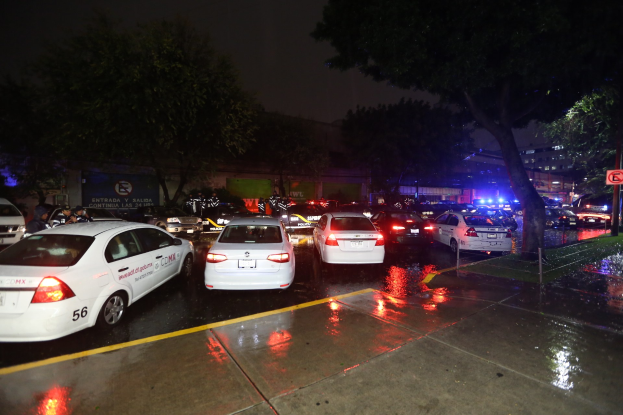 Eine Gruppe von Polizeiwagen auf einem Parkplatz bei Nacht mit sichtbarer Beschilderung, Gras, Bäumen, Gebäuden, einer Fahne, Lichtern und einem bewölkten Himmel.