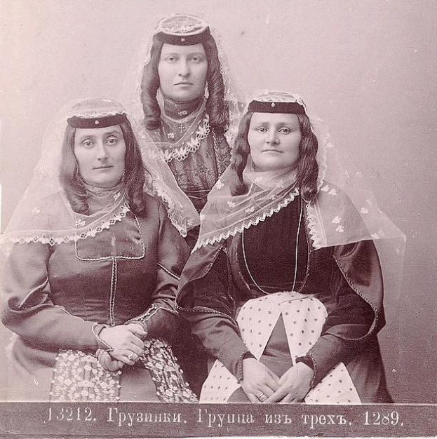 Schwarzes und weißes Foto von drei Frauen in traditionellen russischen Hochzeitskleidern mit farbenfroher Stickerei und Perlen, die vor einer einfarbig weißen Wand stehen.