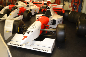 Eine Gruppe von Rennwagen in einem Museum ausgestellt, umgeben von Menschen, ein Schild, eine Absperrung und eine Wand im Hintergrund.