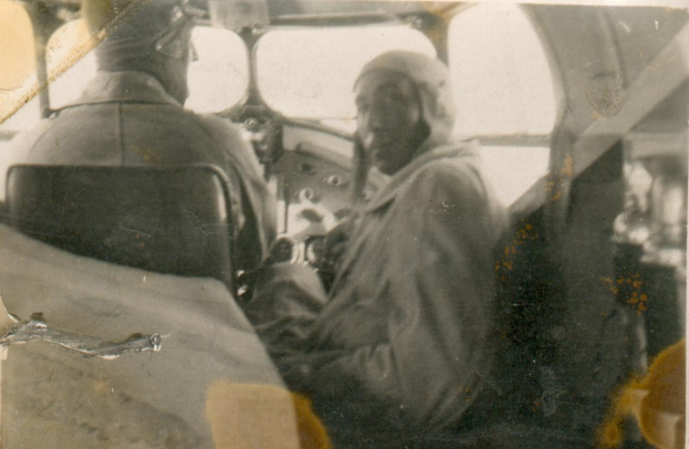 Zwei Männer in Schwarz-Weiß-Foto, einer sitzt im Cockpit und trägt eine Pilotenuniform, der andere steht neben ihm, beide mit ernsten Gesichtern.