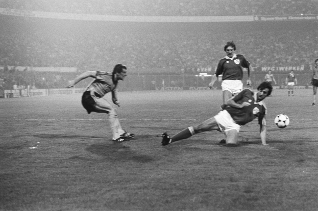 Schwarzes und weißes Foto von Männern, die auf einem Feld Fußball spielen, das von einem Zaun umgeben ist, mit Zuschauern im Hintergrund.