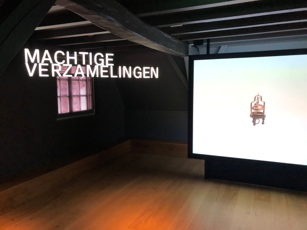 Ausstellungsinstallation 'Machtige Verzamelingen' im Museum of Modern Art in Berlin mit einem großen Bildschirm mit Text an der Wand rechts und einem Fenster links sowie der Decke oben.