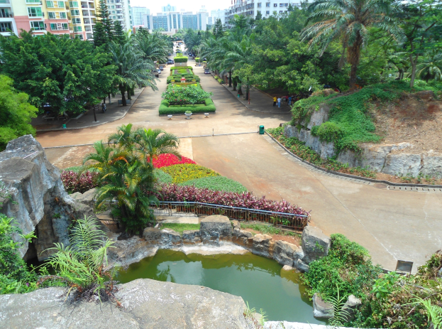 Luftaufnahme eines Parks mit einem Teich, umgeben von Felsen, Pflanzen, Bäumen und Blumen, mit Gebäuden im Hintergrund und einem klaren blauen Himmel, mit Menschen, die herumlaufen.