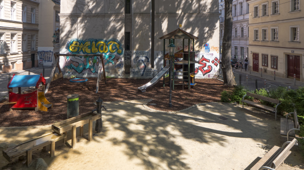 Spielplatz in einer Stadt mit Graffiti, umgeben von Gebäuden, Bäumen, Bänken und einem Müllner, mit Menschen und Fahrzeugen auf der Straße und dem Himmel im Hintergrund.