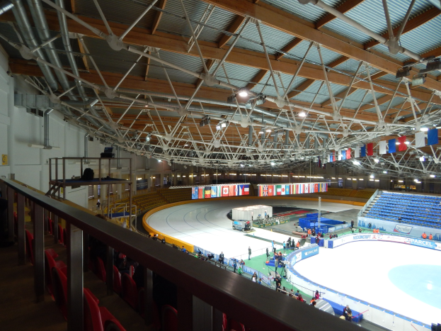 Große Indoor-Eisbahn mit zahlreichen Menschen, die Schlittschuh laufen, umgeben von Sitzplätzen, Geländern, Brettern, Beleuchtungskörpern und konstruktiven Elementen, mit einer Wand im Hintergrund.