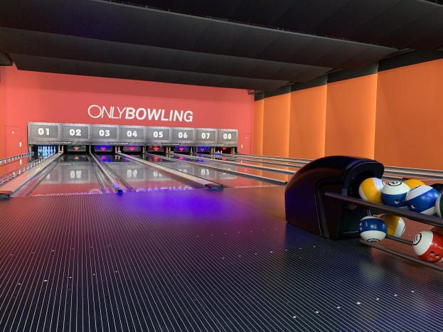 Eine Bowlingbahn mit Bowlingkugeln rechts, Pins in der Mitte, einer textbedeckten Wand im Hintergrund und sichtbarer Decke.