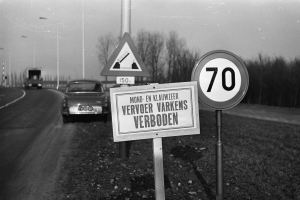 Ein Schwarz-Weiß-Foto eines Tempolimitschildes am Straßenrand mit einem vorbeifahrenden Auto, Bäumen, Pfählen und einem klaren Himmel im Hintergrund.