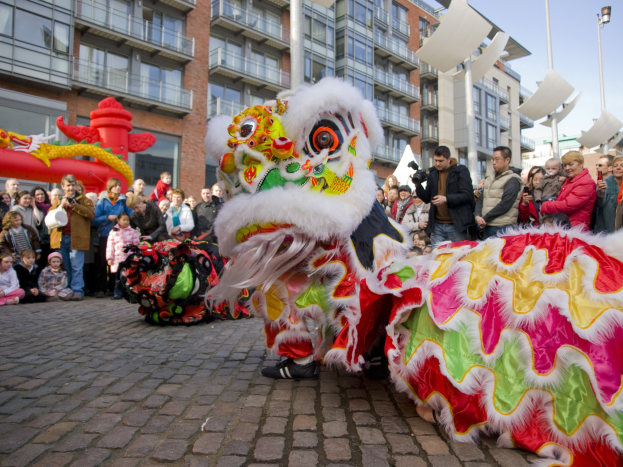 Ein farbenfrohes chinesisches Neujahrsfest in Amsterdam mit einem Löwen tanzen vor einer Zuschauermenge, einige halten Kameras, vor dem Hintergrund von Gebäuden, Laternenmasten und einem klaren blauen Himmel.