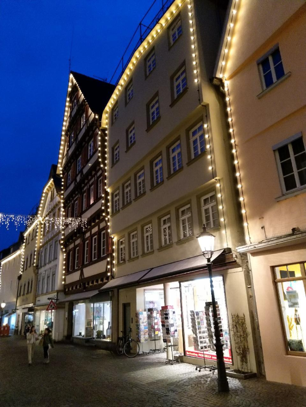 Eine Kopfsteinpflasterstraße in Rothenburg ob der Tauber, Deutschland, beleuchtet von Weihnachtslichtern mit Menschen und einem geparkten Fahrrad, flankiert von Gebäuden mit Fenstern unter einem sichtbaren Himmel.