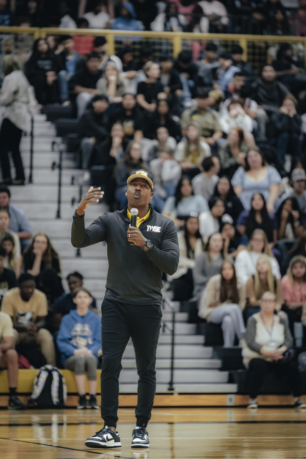 Ein Mann mit Mütze hält ein Mikrofon auf einem Basketballfeld, hinter ihm sitzen Menschen auf einer Treppe und Taschen auf dem Boden.
