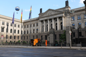 Das Reichstagsgebäude in Berlin, Deutschland, ein großes Gebäude mit zahlreichen Fenstern, umgeben von Straßenmasten, Laternen, Schildern, Fahrzeugen, Fußgängern, Flaggen, Gittern und Bäumen, unter einem bewölkten Himmel.
