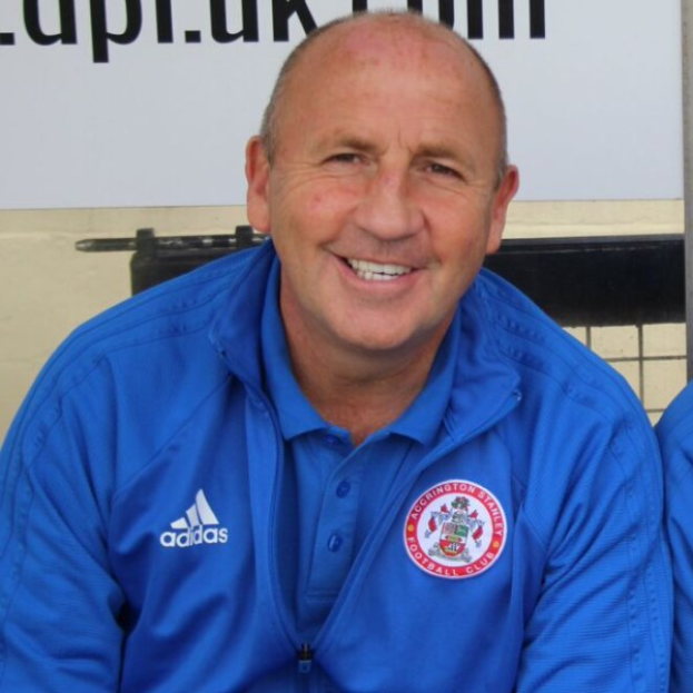 Zwei Männer in einem Raum, einer in einem blauen Jackett neben dem anderen, mit einer Tafel an der Wand dahinter.