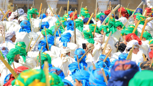 Eine große Gruppe von Menschen in bunten Kostümen marschiert die Straße entlang, jeder hält einen Stock in der Hand und nimmt an einem Festival mit einem Gebäude im Hintergrund teil.