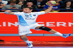 Ein Mann in der Luft, der einen Handball hält, mit einer Gruppe von Menschen im Hintergrund und einem Banner mit Text dahinter.