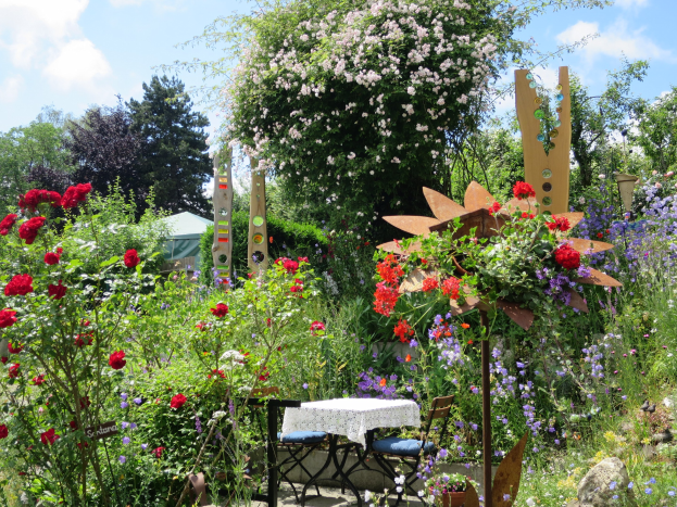 Ein Garten mit bunten Blumen und Pflanzen, der einen Tisch mit einer Decke und Stühlen zeigt, vor Bäumen und einem bewölkten Himmel im Chelsea Flower Show.