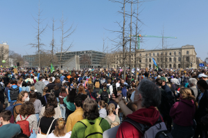 Eine große Menge von Menschen, die an einer Klimademonstration teilnehmen, stehen vor einem Gebäude mit Bäumen und einem klaren Himmel im Hintergrund, viele halten Schilder und tragen Täschchen.