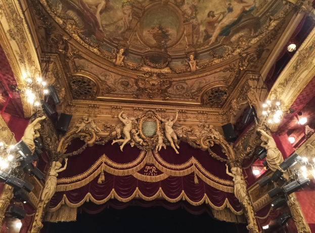 Innere eines Theaters mit einer roten Gardine, Kronleuchtern, Skulpturen, Lichtern und einer Deckenmalerei, die eine Bühne mit ein paar Menschen unten zeigt.