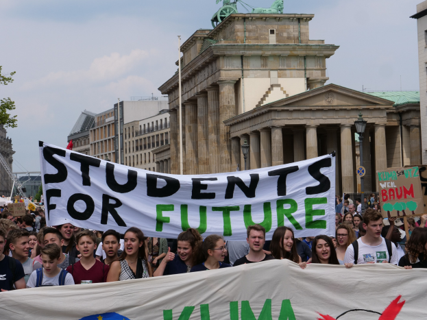 Eine Gruppe von Schülerinnen und Schülern marschiert in Berlin, eine bunt bemalte "Students for Future"-Fahne schwenkend, vor einer Kulisse aus Gebäuden, Bäumen und Himmel.